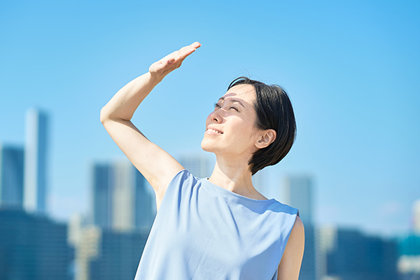 A woman, with a city in the background, looks at the sky, holding her hand in the air to give shade to her eyes.