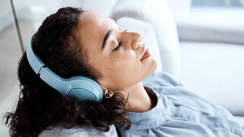 A woman sits on a sofa with her head over the the top of the pillow, eyes closed, and headphones.