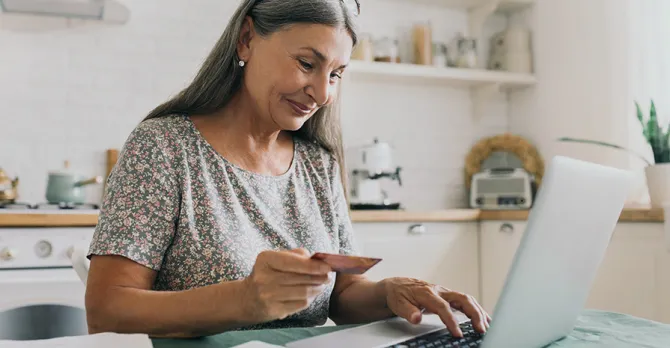 A woman sits at her kitchen table and uses her credit card to pay a bill on her laptop.