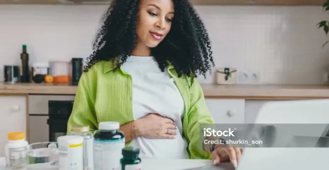A pregnant woman does research on her laptop, which is next to vitamin bottles.