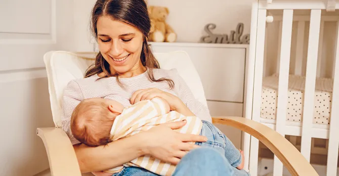 A mother sits in a chair in the baby's room while breastfeeding.