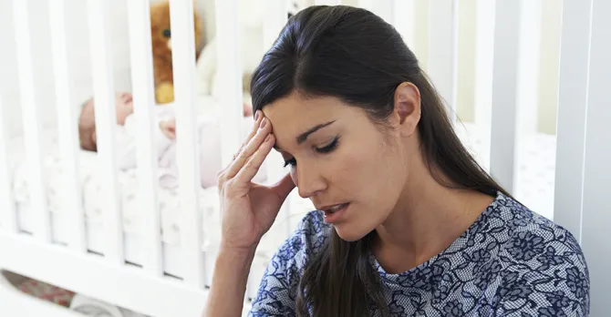 A new mother sits on the floor, just outside of the baby's crib.  Part of her hand covers part of her face, and her fingers are on her temples.