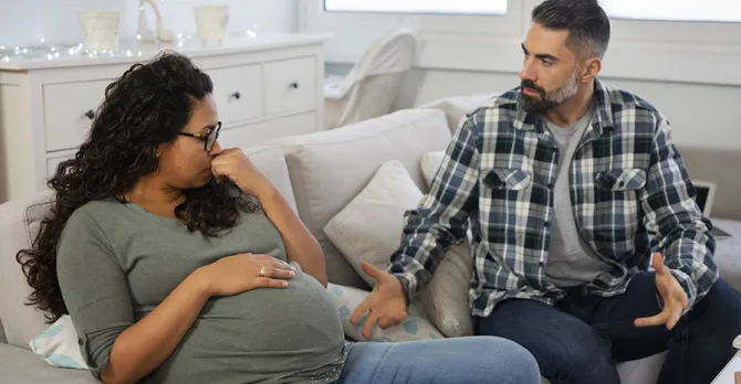 Expecting parents on a sofa.  The pregnant woman is not pointing toward the man, but she looks to the side at him and she looks sad.