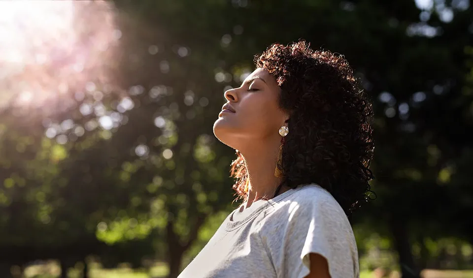 A woman stands with her eyes closed and head tilted back, being mindful and admiring the sun shining through the trees above her.