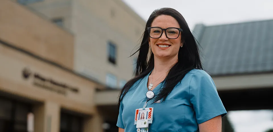 A smiling female nurse in blue scrubs is standing in front of Marlborough Hospital. 