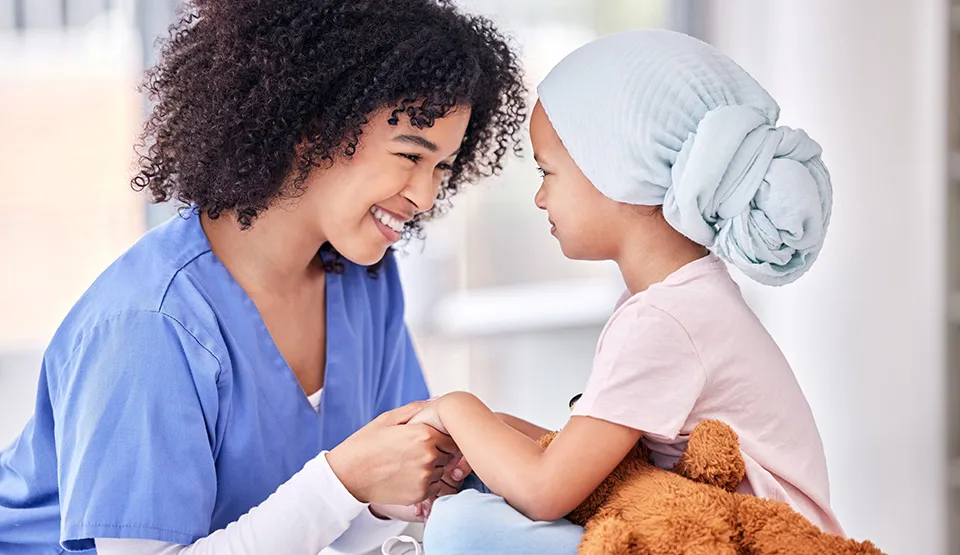 A smiling caregiver holds the hands of a young patient wearing a head wrap.