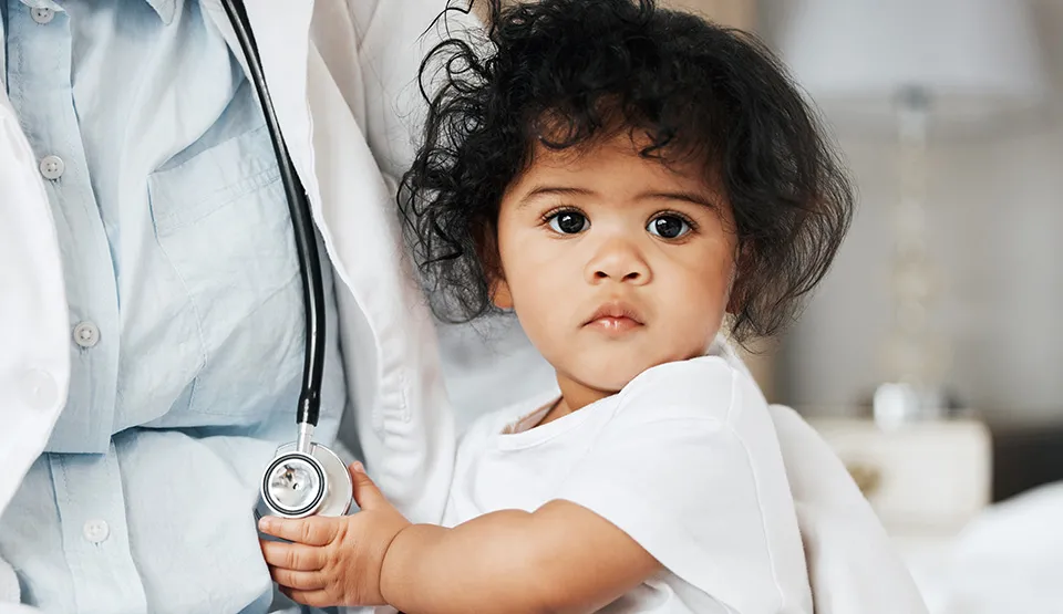 A young child in the arms of a provider, holding a stethoscope and looking at the camera.