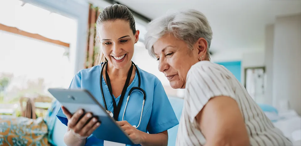 A caregiver visits a patient in her home and both look at the information on the caregivers tablet.