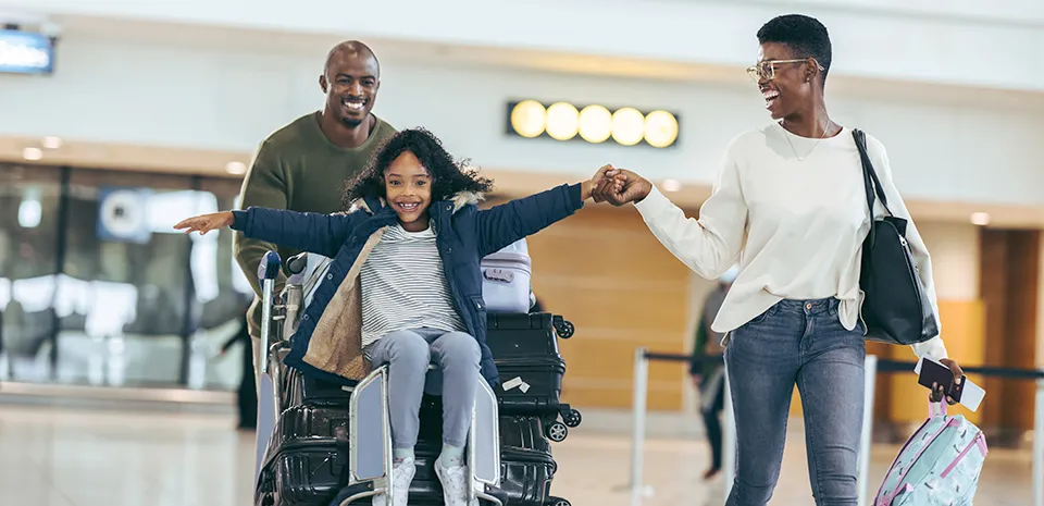 A family at an airport, with a child riding on the stack of suitcases, preparing for travel together.