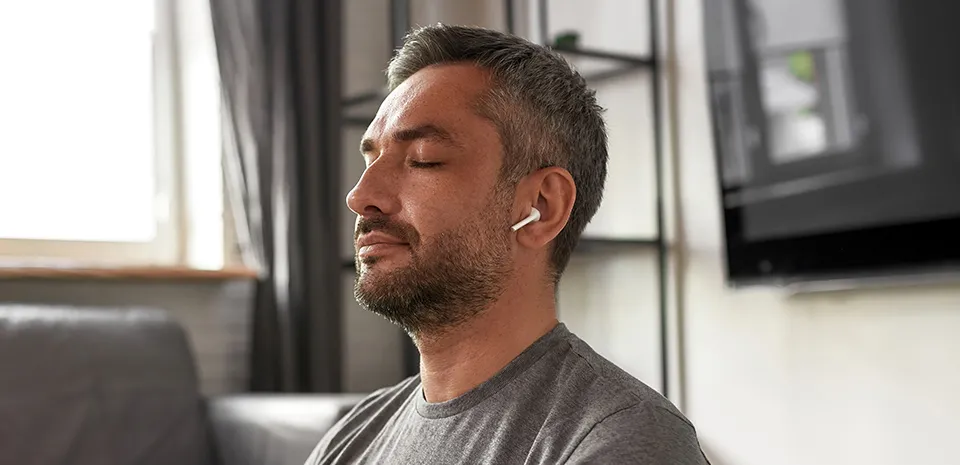 A man sits on a floor with correct posture. He is wearing earbuds and has his eyes closed.