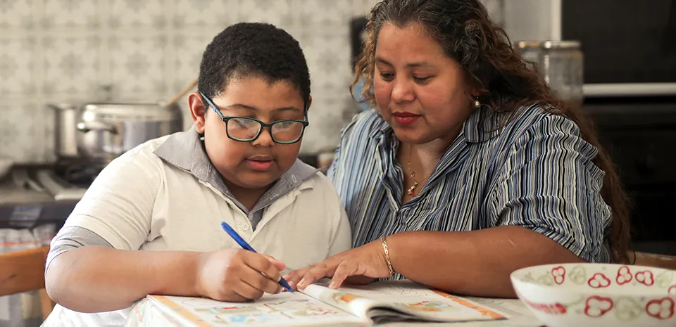 An adult helps a young boy with his homework at the kitchen table.