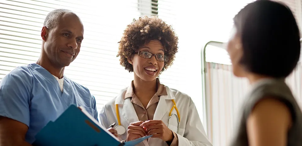 A male and female caregiver speak to a woman in an exam room setting. 