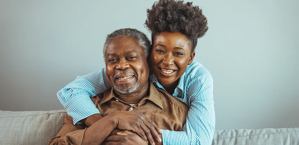 A man sits on a couch, looking at the camera, smiling, while a woman stands behind him, wrapping her arms around him.