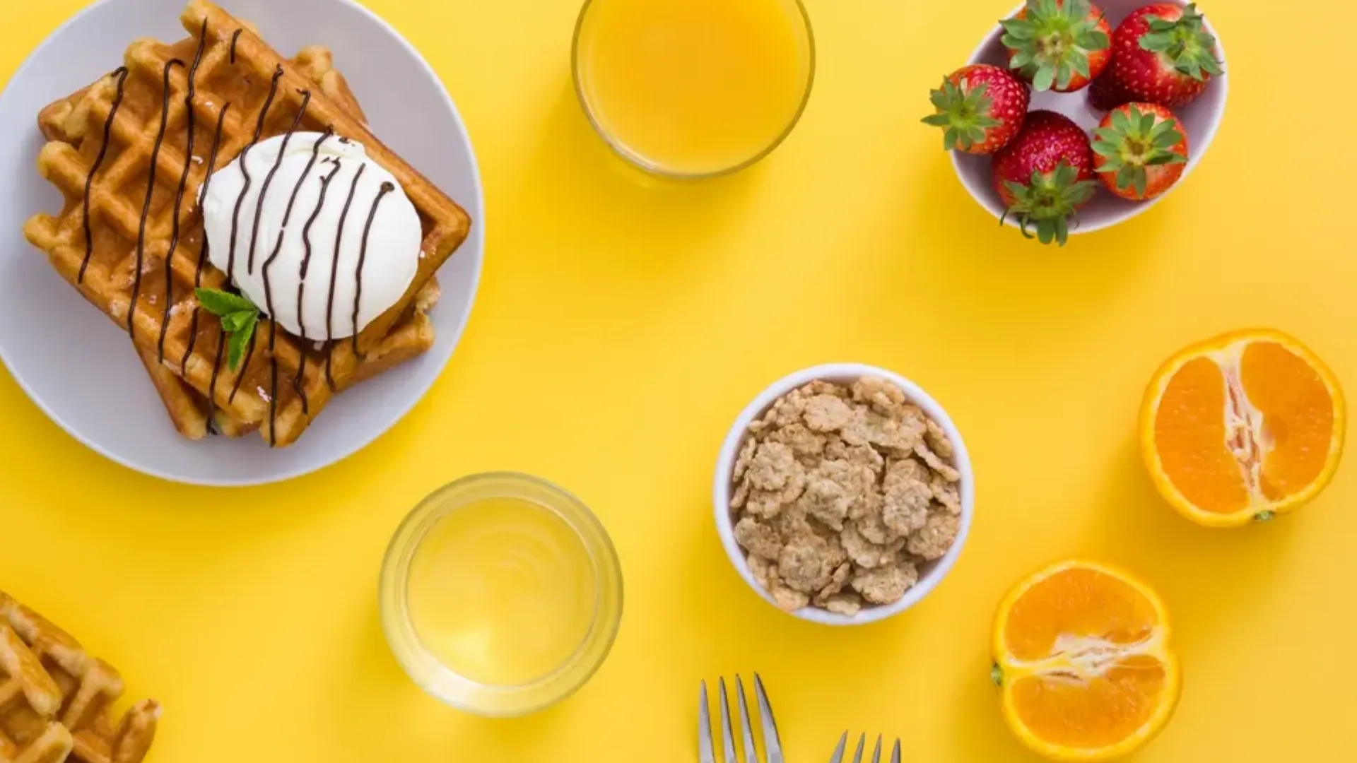 Waffles, cereal, fruit, and juice are shown from above on a yellow table.