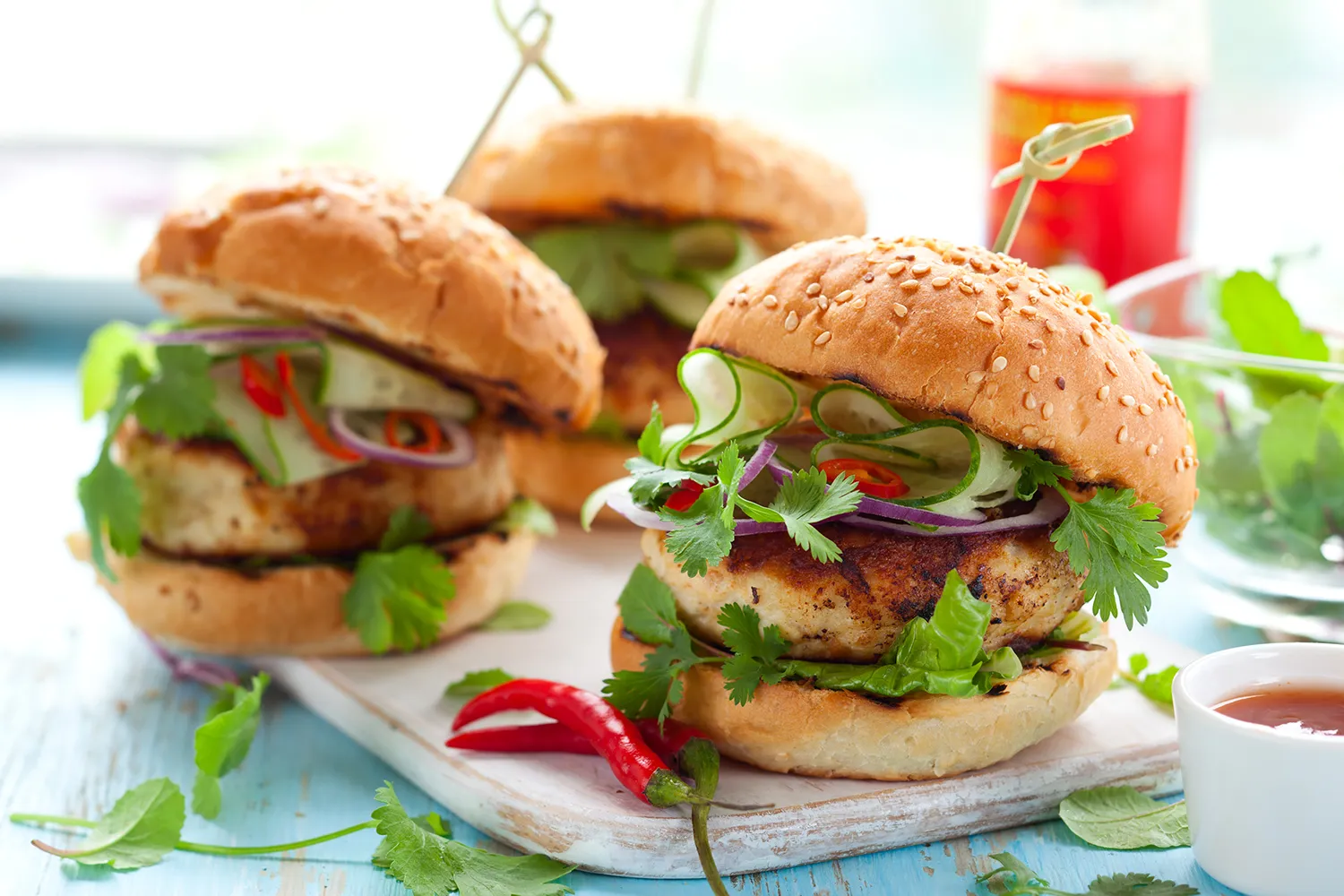 Several turkey burgers with various vegetables on sesame seed buns are shown on a cutting board.