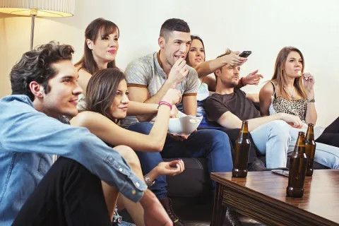 A college age group of teens are drinking and watching tv.