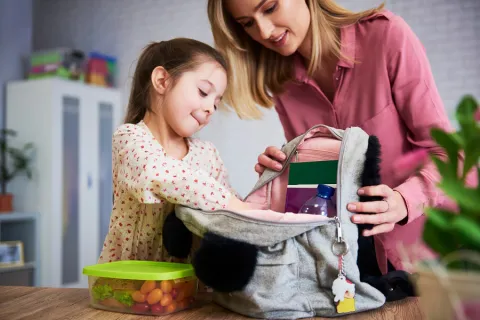 A young girl puts things into her backpack while her mother holds it open on a countertop.