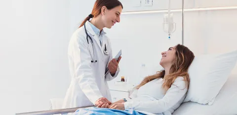 A female doctor standing next to a patient's bed. The doctor is smiling at the female patient, who is also smiling at the doctor.