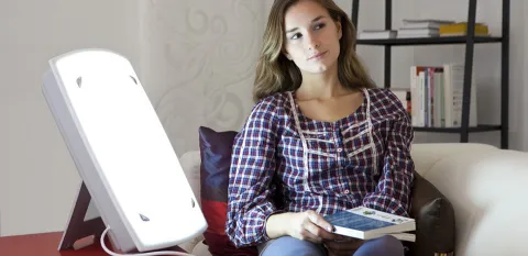 A young woman sits on a sofa near the light used to treat her seasonal affective disorder.