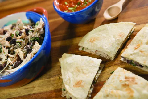 A blue bowl of shredded chicken and black beans is next to a quesadilla that is cut into quarters.