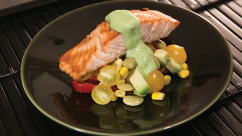 A piece of salmon with a green avocado sauce is next to a cherry tomato and corn salad, all on a black plate.
