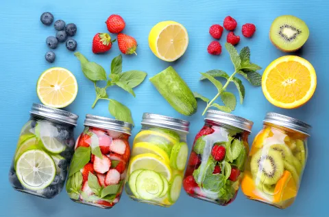 Multiple mason jars on a blue background have fruits and vegetables spilling out.