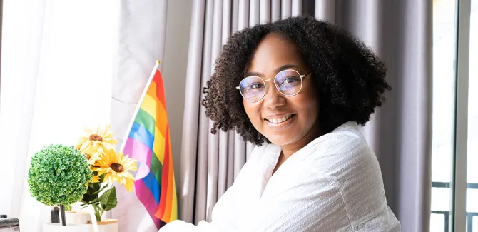 A black woman, smiling at a desk that has flowers and a pride flag displayed on it.