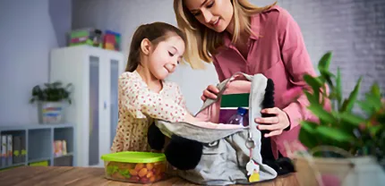 A young girl puts things into her backpack while her mother holds it open on a countertop.