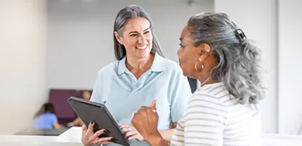 A woman with a tablet speaks to another woman in a hospital setting.