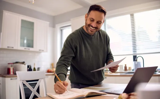 Man at kitchen table writing goals in a book with laptop open