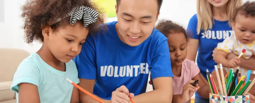 Young man volunteering at a daycare helping a young girl draw.