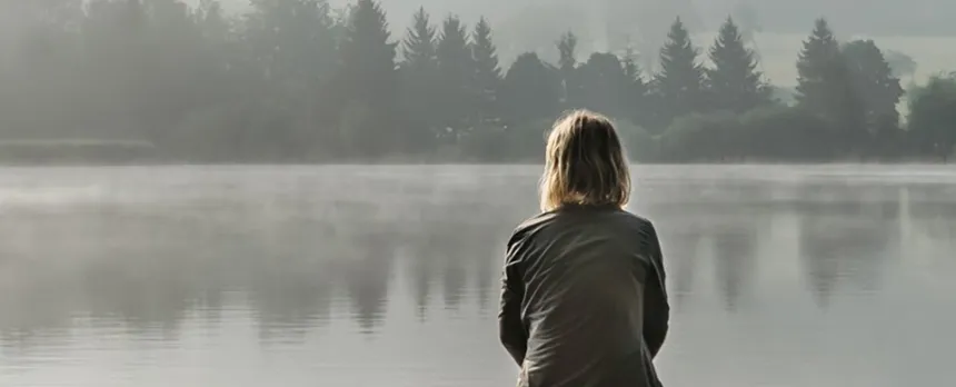 A woman sits on a dock, looking across the water through a thick fog.