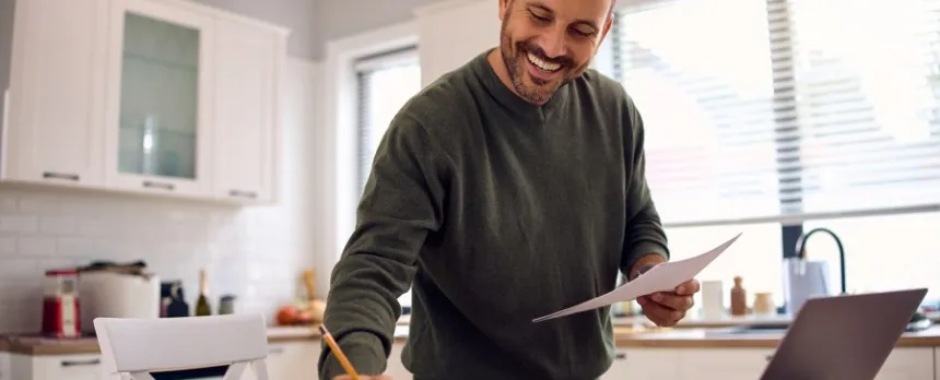 Man at kitchen table writing goals in a book with laptop open