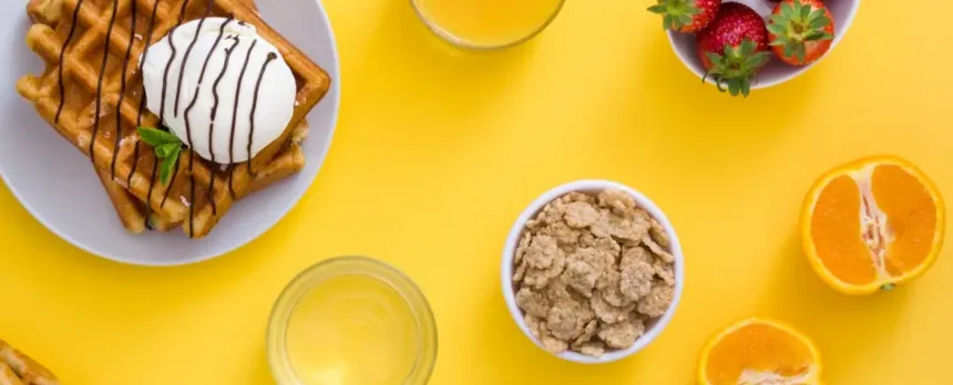 Waffles, cereal, fruit, and juice are shown from above on a yellow table.