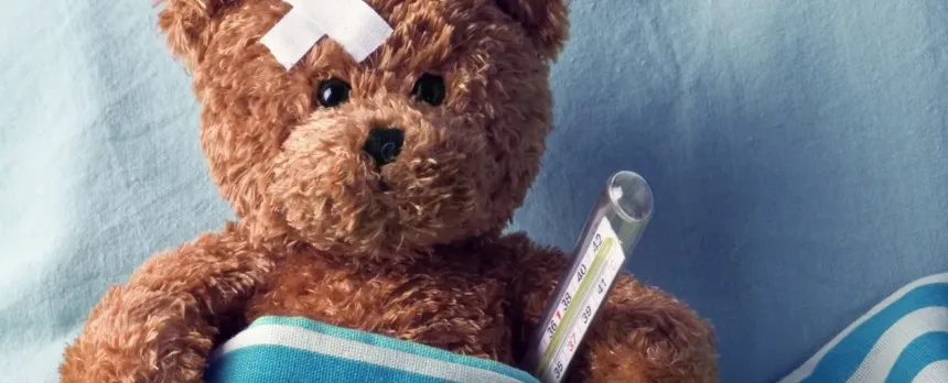 A stuffed teddy bear is in a hospital blanket with a thermometer and bandage on its head.
