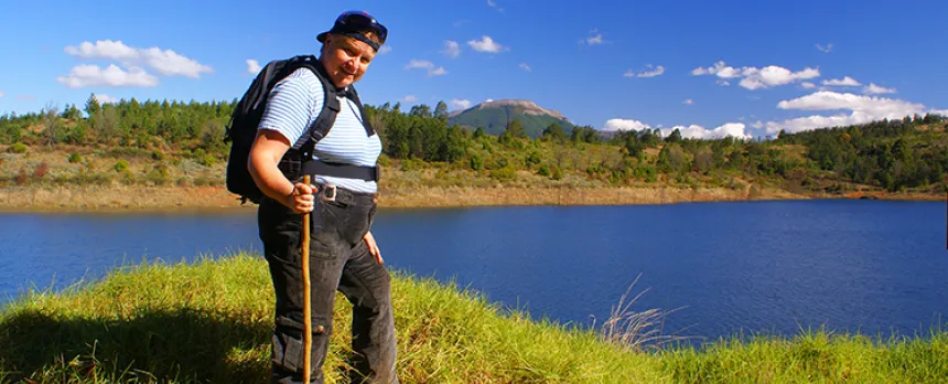 A mature woman is hiking and posed in front of a lake and mountains.