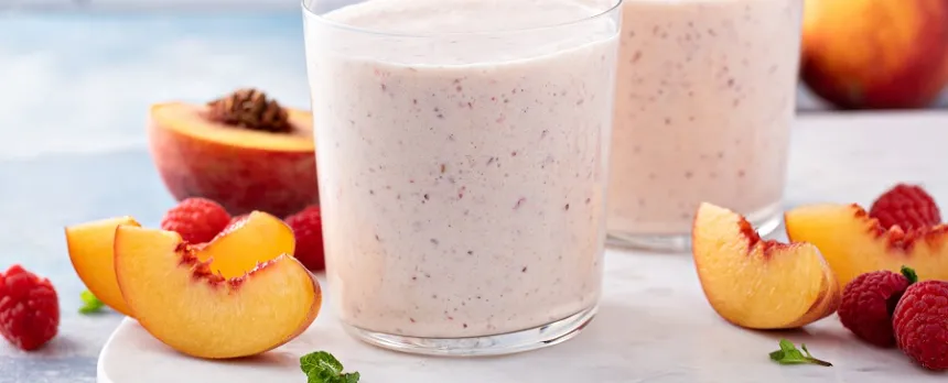 Two smoothie glasses are surrounded by peaches and other berries.