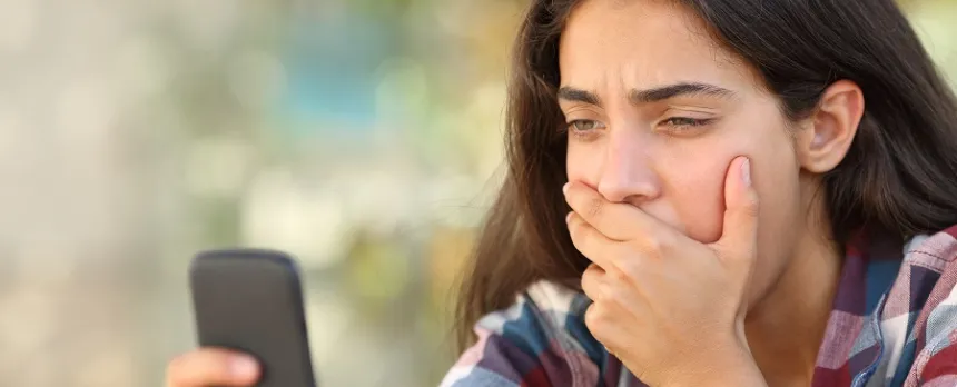 A teen is covering their mouth while looking at a cell phone.