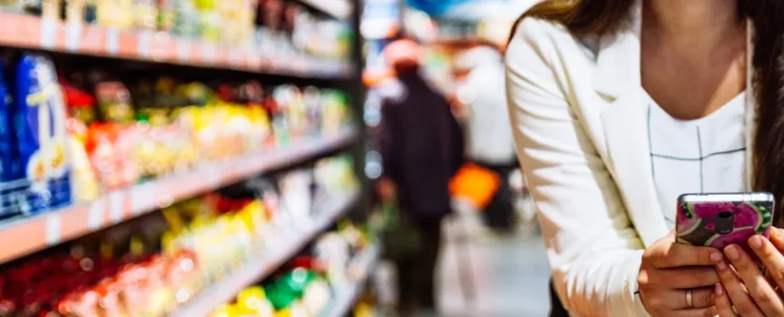 A closeup shows a woman on her cell phone shopping in a grocery store.