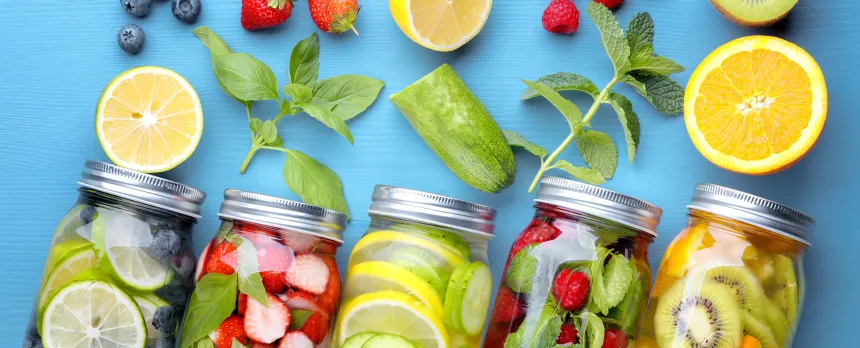 Multiple mason jars on a blue background have fruits and vegetables spilling out.